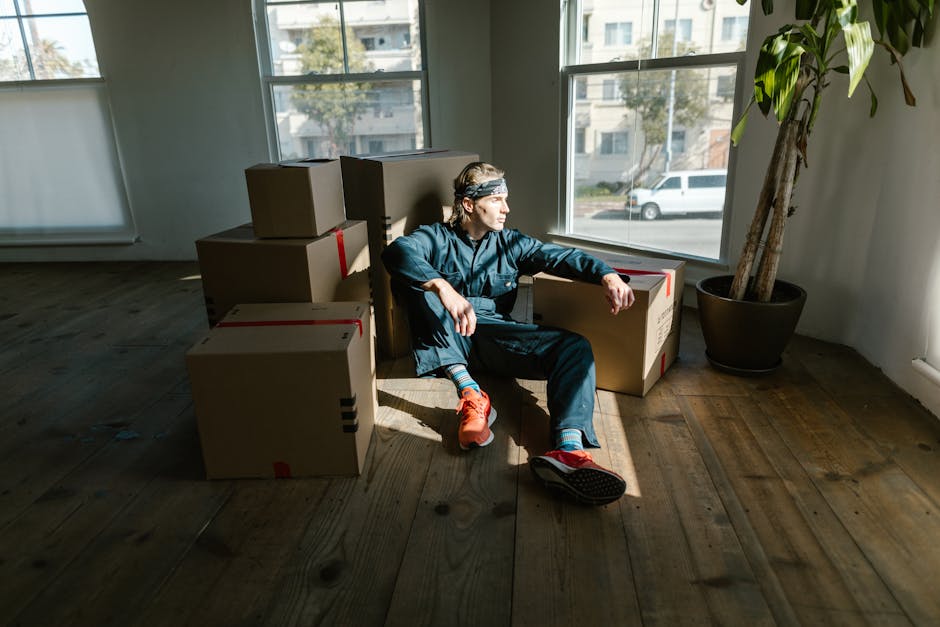 A person wearing a blue jumpsuit, red sneakers, and a headband is sitting on a wooden floor inside a room, surrounded by stacked cardboard moving boxes of various sizes. The individual is leaning back against the boxes with one arm resting on a box and the other on their knee, gazing out of large, sunlit windows that reveal a residential street with parked cars and buildings outside. A large potted plant with green leaves is positioned in the corner near the window. This scene suggests a home relocation process, with boxes prepared for packing or unpacking during a furniture transport or moving logistics activity. The presence of packing materials and the arrangement of boxes imply the ongoing packing and moving operations carried out by [COMPANY_NAME], such as at the initial stages of a house removal or apartment move. The natural lighting and minimal furnishings create a neutral, professional setting focused on the unpacking or packing phase of the move.