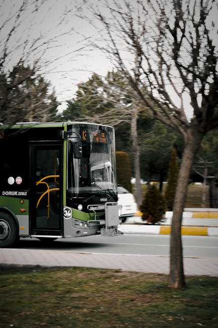 A black and green public bus is seen parked alongside a street with leafless trees lining the pavement. The bus displays route information on an electronic sign above the windshield and is part of a moveable fleet used for local transportation. In front of the bus, there is a trolley with a white box attached to a trolley app, indicating loading or unloading activity. Nearby, a white car is partially visible behind the trees. The scene takes place during daytime with overcast weather, and the background includes additional vehicles and residential or commercial buildings. This setting illustrates urban logistics involved in home relocation or furniture transport, supported by professional moving services such as Man with Van Stepney, utilised for packing and loading processes as part of a comprehensive removals operation.