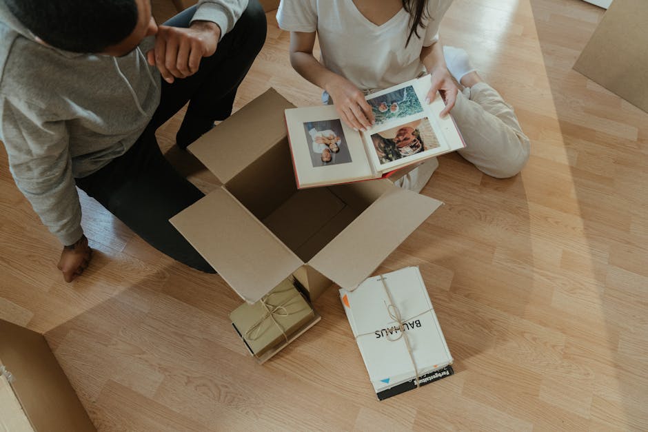 A person wearing a blue jumpsuit, red sneakers, and a headband is sitting on a wooden floor inside a room, surrounded by stacked cardboard moving boxes of various sizes. The individual is leaning back against the boxes with one arm resting on a box and the other on their knee, gazing out of large, sunlit windows that reveal a residential street with parked cars and buildings outside. A large potted plant with green leaves is positioned in the corner near the window. This scene suggests a home relocation process, with boxes prepared for packing or unpacking during a furniture transport or moving logistics activity. The presence of packing materials and the arrangement of boxes imply the ongoing packing and moving operations carried out by [COMPANY_NAME], such as at the initial stages of a house removal or apartment move. The natural lighting and minimal furnishings create a neutral, professional setting focused on the unpacking or packing phase of the move.