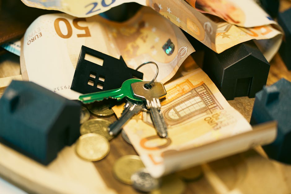 Close-up image showing keys, including a house key with a green plastic head and a metal keyring, resting on a pile of assorted euro banknotes and coins, with packing materials and a small black house-shaped keychain nearby. The scene appears to be inside a property, possibly during a home relocation process managed by Man with Van Stepney, with some cardboard and fabric wrapped items visible, and the arrangement indicating packing and moving activities associated with furniture transport and household removals.