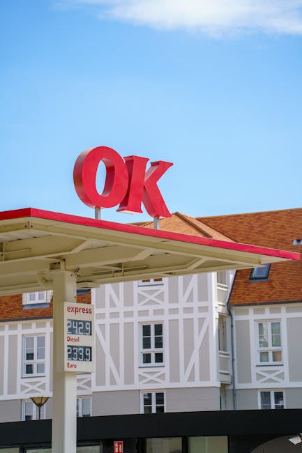 Photograph captures the exterior of a residential property with a white and grey facade, featuring a gabled roof with red tiles and multiple windows. In the foreground, a petrol station canopy with a red trim extends outward, supported by white columns. Prominently mounted on top of the canopy is a large, red, three-dimensional sign displaying the letters 'OK'. Underneath the canopy, there are large digital signage boards showing fuel prices for express, diesel, and Euro fuels, with costs listed in pence and euros. The setting appears to be a clear day with a bright blue sky. This scene illustrates a typical urban environment where furniture or belongings might be loaded or unloaded during a house removal or relocation process, with the petrol station potentially serving as a stop during a home-moving service provided by companies like Man with Van Stepney.