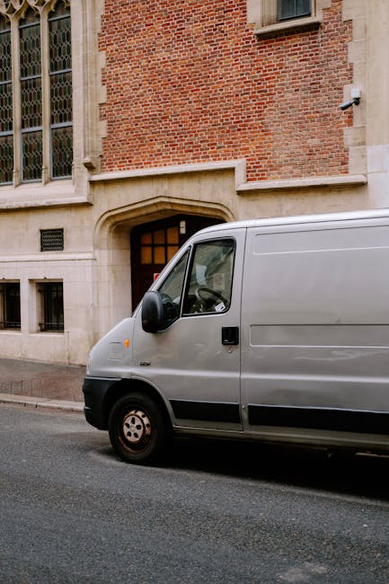 A silver cargo van parked on a city street in front of a historic brick building with large stained-glass windows. The van is positioned close to a stone doorway, indicating it is in the process of loading or unloading furniture or moving boxes as part of a home relocation. The side door of the van is closed, and the vehicle is on an asphalt pavement with no visible occupants. This scene captures the loading process typical of professional removals services provided by Man with Van Stepney, with the focus on efficient transport and logistics for local house moves. The lighting is natural, and the environment appears calm, with no other vehicles or pedestrians visible in the image.