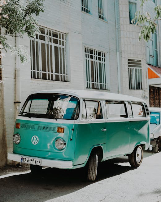 A white vintage van parked on a residential street with a suburban house in the background, featuring a brick exterior and a chimney. The van is positioned next to a grassy area separated by short wooden posts, and it appears to be used for house removals or furniture transport. The van’s side doors are closed, and the vehicle is clean but shows signs of age. Several tall trees with green and brown leaves surround the scene, indicating mild weather and daylight conditions. This image illustrates a home relocation process, with the van ready for loading or unloading goods, possibly part of a professional removals service such as Man with Van Stepney, supporting packing, moving, and loading activities in the context of same-day house moves.