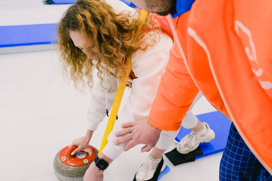A woman with curly red hair wearing a white hoodie and white sneakers is seen in a home interior, positioning a large rock climbing hold or similar object on the floor, aided by a man in an orange jacket and blue checkered trousers. The woman is kneeling on a blue foam mat, leaning forward with an intense focus, while the man supports her by holding her by the back and standing close beside her. Both are participating in a moving or packing activity related to house removals, with the indoors illuminated by bright lighting, and various packing materials or equipment possibly out of frame. This scene highlights careful handling and positioning of household items during home relocation, reflecting the services of companies like Man with Van Stepney, which facilitate furniture transport and packing for house moves.