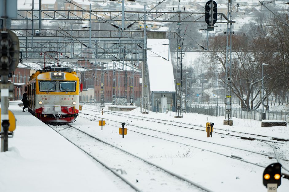 A yellow and red electric train approaching a snow-covered railway station platform during winter daytime, with overhead electric lines and metal structures spanning across the tracks. The platform has some scattered snow, and a person is seen near the train. In the background, there are leafless trees, buildings, and a cloudy sky. This scene illustrates a typical home relocation environment where careful planning for property moving logistics, including transport via train, can be essential during winter months. Man with Van Stepney specialises in removals and packing services, ensuring smooth furniture transport and loading processes even in adverse weather conditions, highlighting their expertise in managing house removals during winter for clients preparing for moves like those outlined in the 'Preparing a Stepney Move for Winter 2026' guide.