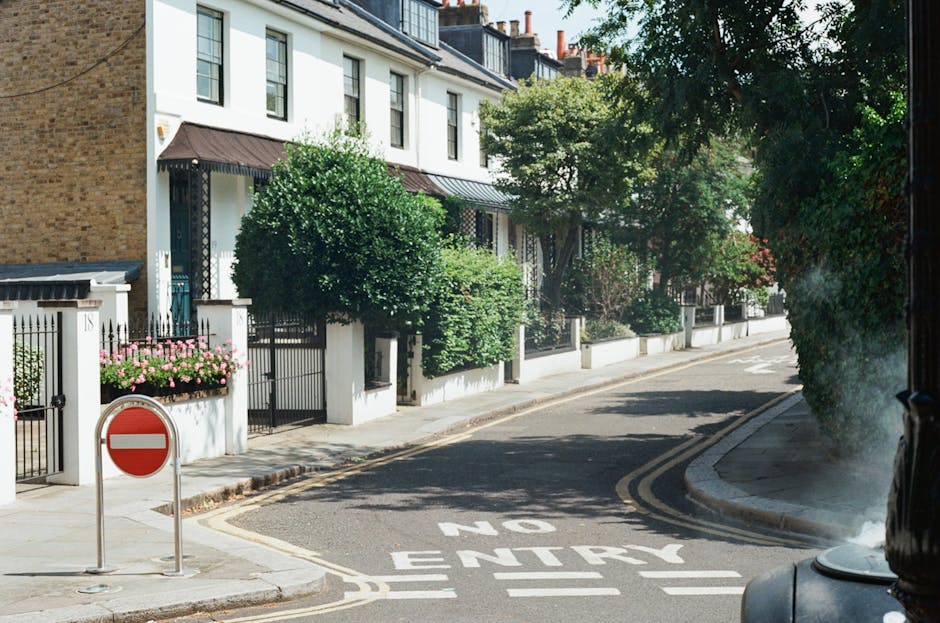 A quiet residential street with a white multi-storey house featuring black window frames, situated behind a white boundary wall decorated with pink flowering plants and lush green trees and bushes. The street has double yellow lines, and a 'No Entry' traffic sign is installed on the pavement near the entrance to a restricted area. The roadway is marked with white painted text indicating 'NO ENTRY' at the turn, and the surface appears clean and well-maintained. Adjacent to the curb, a black van is partially visible with the side door open, suggesting a home relocation or furniture transport process in progress. The environment is sunny with clear lighting, and the scene conveys an organized setting suitable for professional removals, such as those provided by Man with Van Stepney, assisting with house removals and moving logistics along Mile End Road.