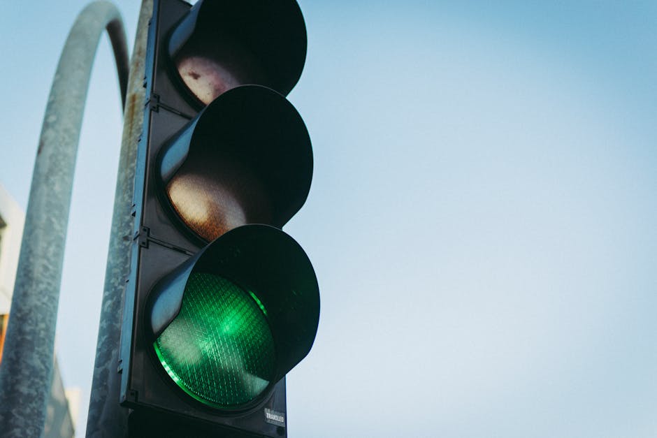A close-up view of a pedestrian traffic light showing a green signal, mounted on a metal pole beside Mile End Road in Stepney. The traffic light features three circular sections: the top two are off with dark lenses, while the bottom section illuminates with a bright green light indicating it is safe to proceed. The background shows a clear, pale blue sky with no visible buildings or other street fixtures. The setting provides context for vehicle and pedestrian movement, relevant to house removals and moving logistics in the area, with Man with Van Stepney assisting with the navigation of local road restrictions during a home relocation project.