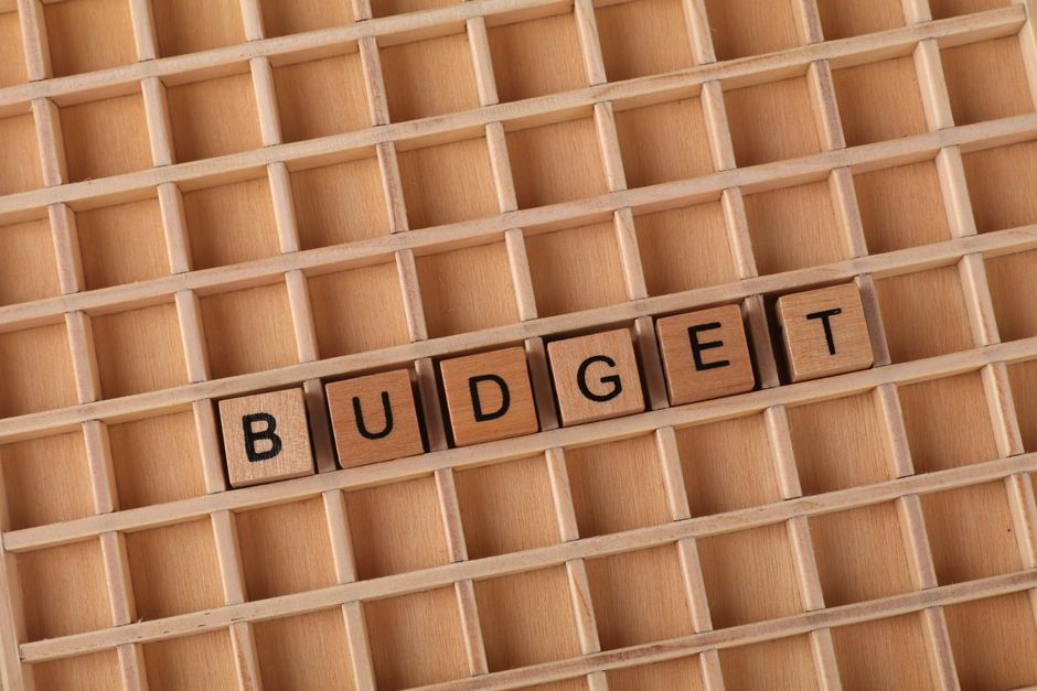 A wooden grid storage tray with multiple small compartments, predominantly empty, with six wooden alphabet blocks spelling out the word 'BUDGET' arranged in a straight line across the middle section of the tray. The tray is placed indoors, likely on a flat surface such as a table or countertop, with natural or artificial lighting illuminating the scene evenly. This image represents the concept of cost considerations or planning within the context of house removals and relocation services, as provided by Man with Van Stepney, supporting topics related to moving logistics, packing, and budget management during home relocation or furniture transport tasks.