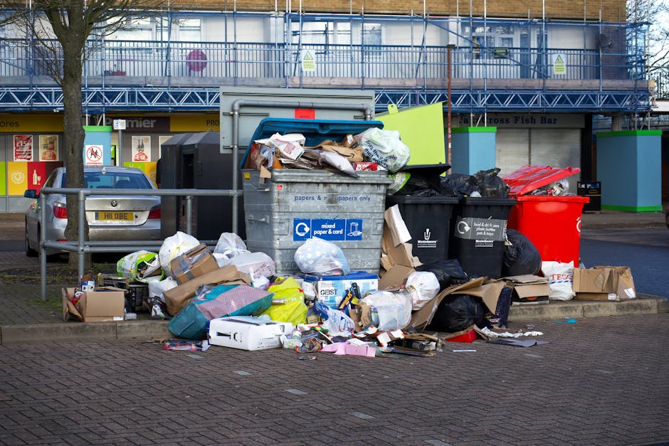 A collection of overflowing waste and recycling bins situated on a paved sidewalk in front of a commercial building in an urban area. The grey mixed paper and card bin is open with its lid tilted back, revealing crumpled papers, cardboard boxes, and other recyclables inside. Surrounding the bin are numerous discarded items, including flattened cardboard boxes, plastic bags filled with rubbish, and loose packaging materials scattered on the ground. There are three additional bins: a black bin for general waste and a red bin for other waste, both filled with black rubbish bags, and a bright green bin with its lid closed. In the background, a parked silver car is partially visible behind a metal railing, with a tree on the left side. The storefront behind displays signage and window displays, and an upper level of the building has scaffolding and protective sheeting, indicating ongoing construction or maintenance. The scene captures a typical urban waste disposal area during the day, before scheduled clearance, highlighting the importance of waste management in house and commercial relocations, as managed by services like Man with Van Stepney.