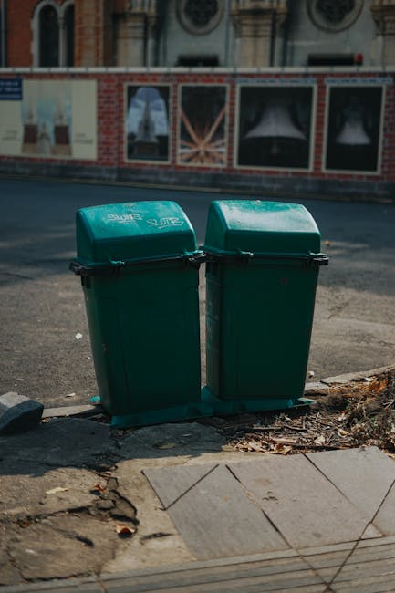 Two green wheelie bins, made of plastic with closed lids, are positioned side by side on the pavement at the edge of a street. They are placed on a small area of uneven ground with dirt and debris nearby, adjacent to a concrete sidewalk with visible cracks and a wooden panel. In the background, there is a building with large windows framed by red brickwork and decorative stone details, suggesting an urban residential or commercial area. The scene is captured in natural daylight, with shadows cast on the pavement. This outdoor setting relates to house removals and moving logistics, as waste disposal and clearance are often part of relocation services provided by companies like Man with Van Stepney. The image reflects the preparation or post-move waste management process involved in home relocation and furniture transport.